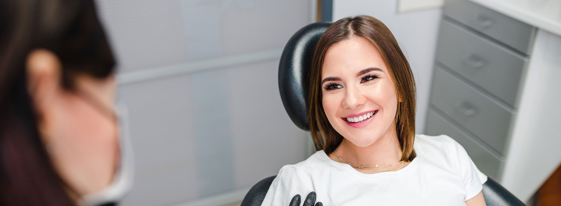 A woman sitting in a dental chair, smiling at the camera, with a dental professional visible behind her.