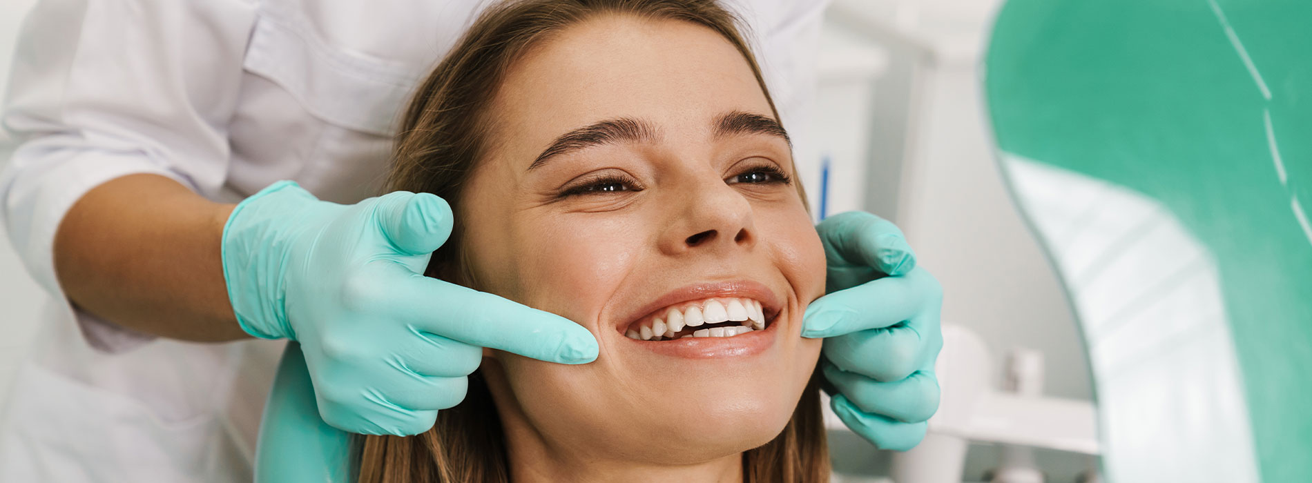 A woman is seated in a dental chair, smiling broadly as a dentist adjusts her teeth with a mirror and tweezers.