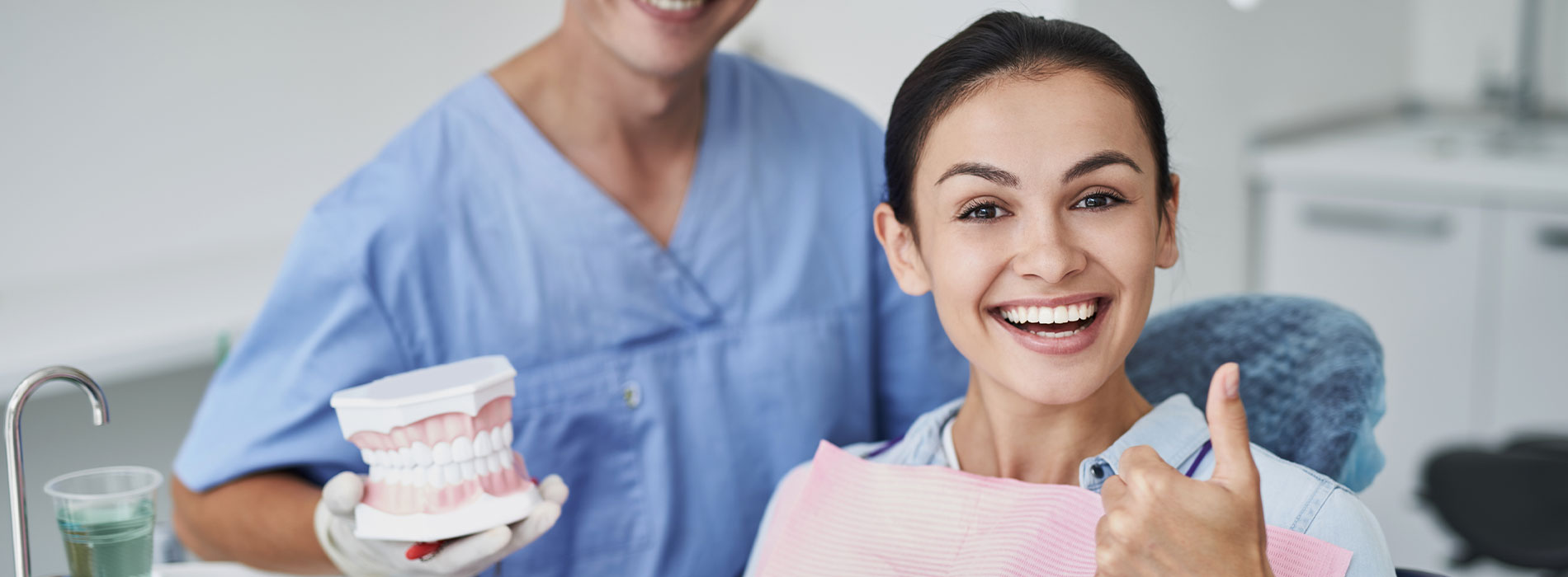 A dental professional in scrubs and a surgical mask, smiling at the camera, with a patient seated next to him, both standing in front of a sink.