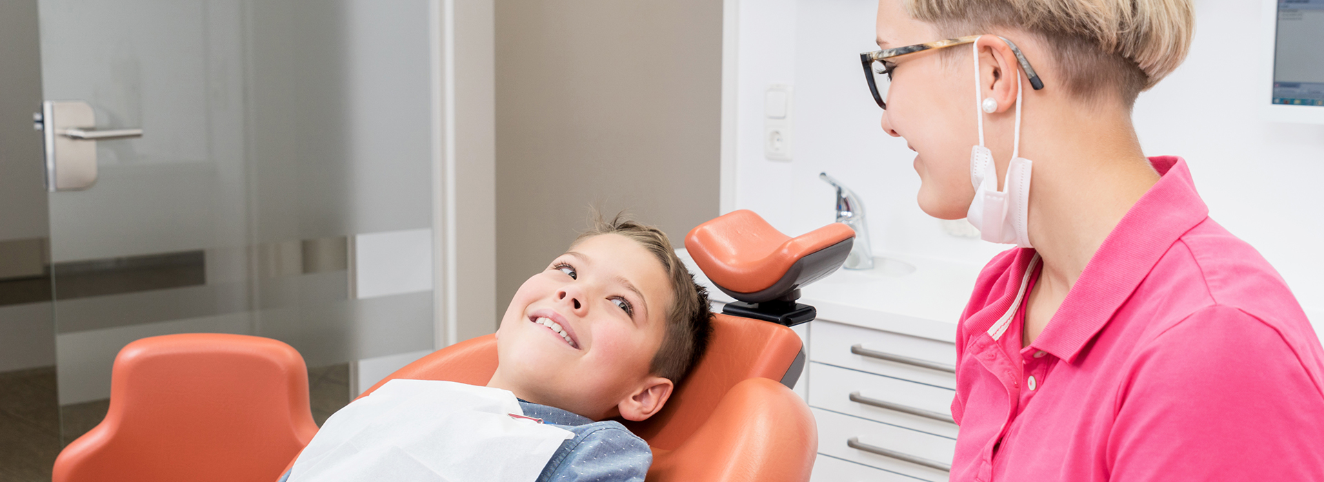 A dentist and a young patient in a dental office, with the dentist seated and smiling at the camera.