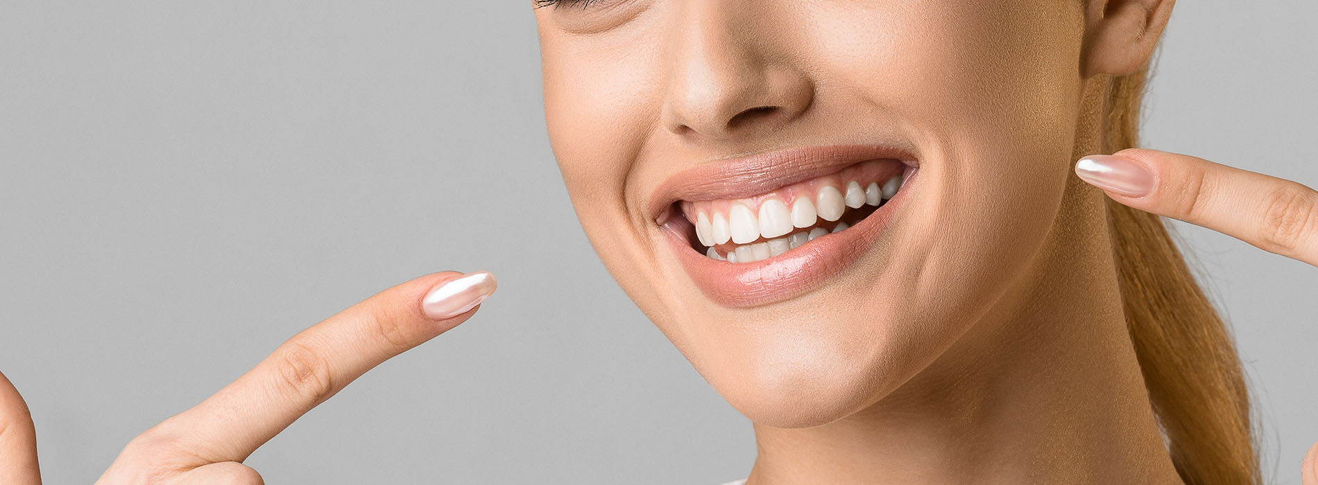 A smiling woman displaying her teeth with a hand gesture, against a neutral background.