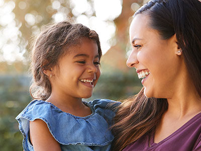 The image is a photograph featuring a woman and a young child, both smiling at the camera. They appear to be outdoors during daylight with natural lighting on their faces.