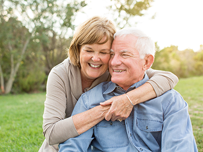 The image depicts an older couple, a man and a woman, embracing each other outdoors during the day.