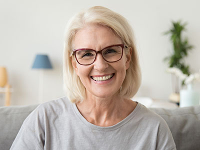 The image shows a woman with short blonde hair, wearing glasses and a light-colored top, smiling at the camera. She appears to be indoors, possibly in a living room setting, with a white wall and a lamp visible behind her.