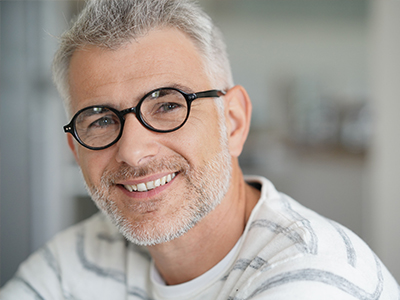 The image shows a man smiling at the camera, wearing glasses and a white shirt with a patterned collar. He has short gray hair and appears to be middle-aged.