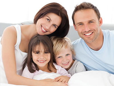 A family of four, including a man, woman, and two children, smiling together in bed.