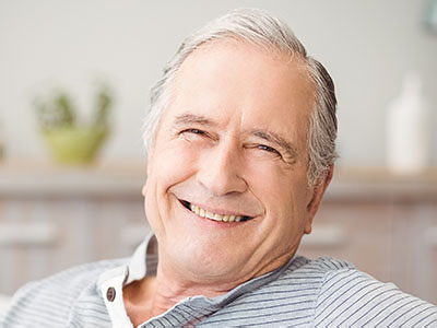 The image shows a smiling older man with gray hair, wearing a blue shirt, seated comfortably in an indoor setting.