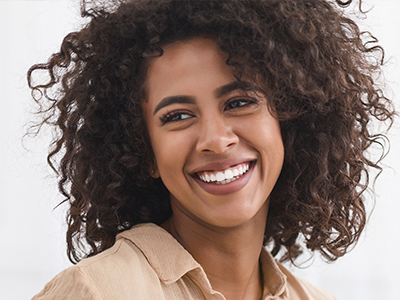 The image shows a woman with curly hair, smiling, and looking directly at the camera. She has a radiant smile and appears to be in a good mood.